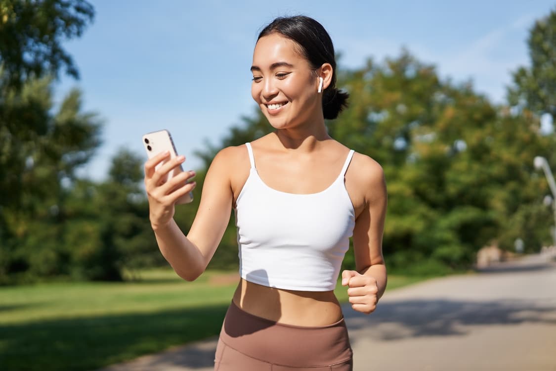 Uma mulher correndo em um parque, olhando para o celular enquanto usa um aplicativo de assessoria de corrida.