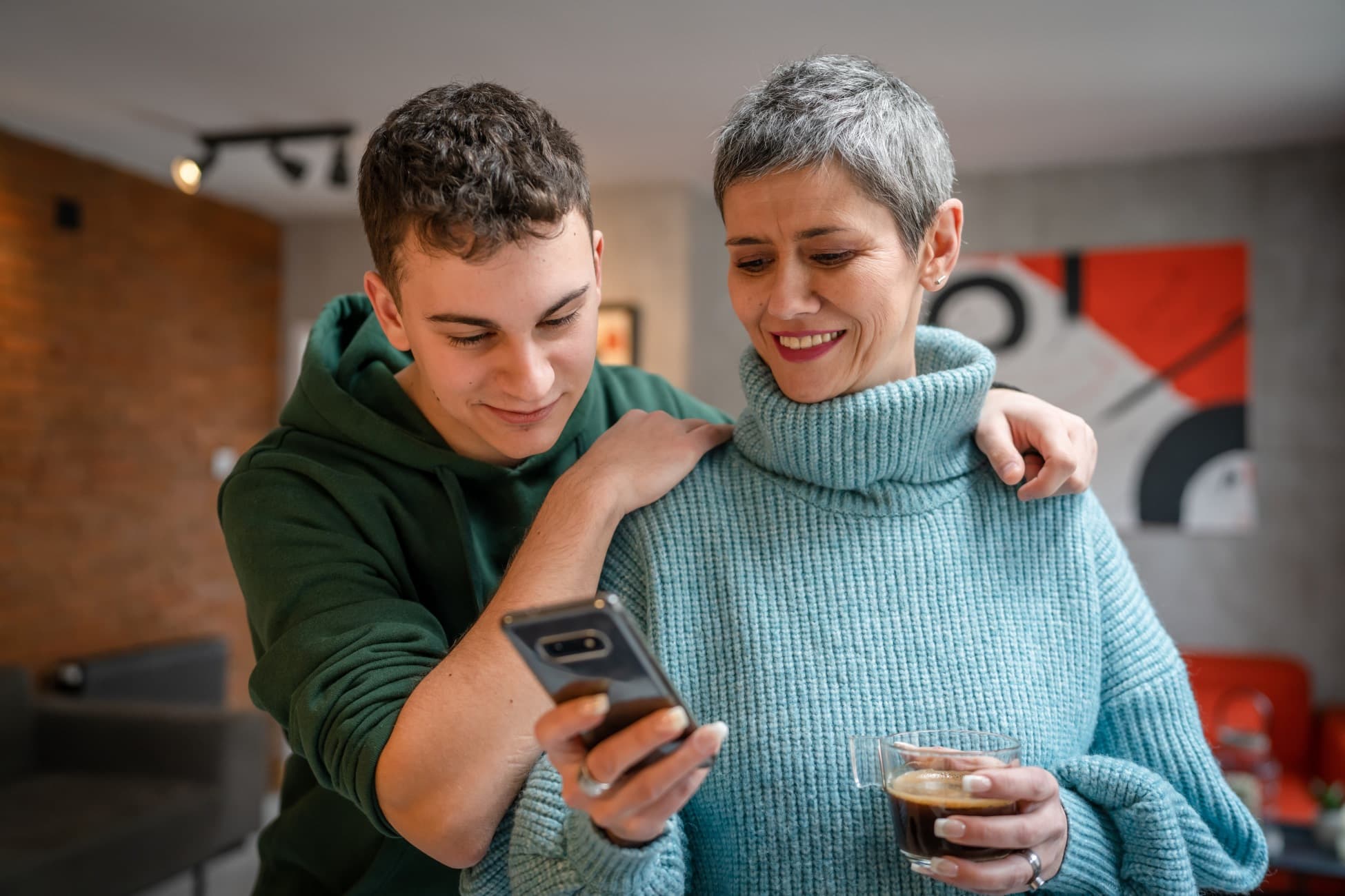 Jovem e mulher de cabelo grisalho sorriem enquanto olham juntos para um celular, em um ambiente acolhedor; eles demonstram confiança ao acessar facilmente o FAQ com perguntas frequentes sobre o tratamento de seus dados pessoais no Hospital Israelita Albert Einstein.