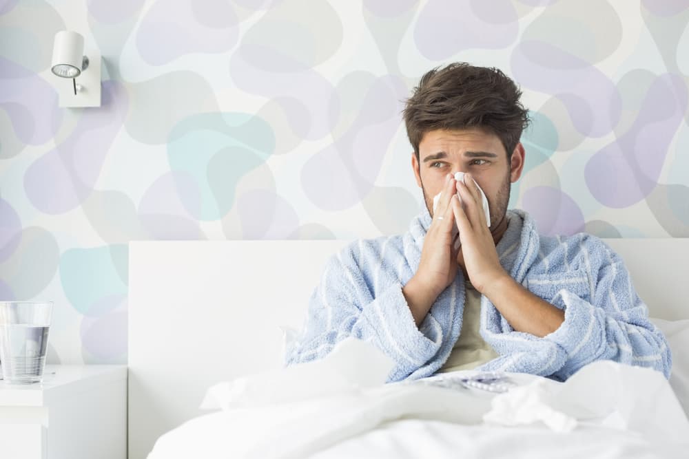 Sick,Man,Blowing,His,Nose,While,Sitting,On,Bed,At