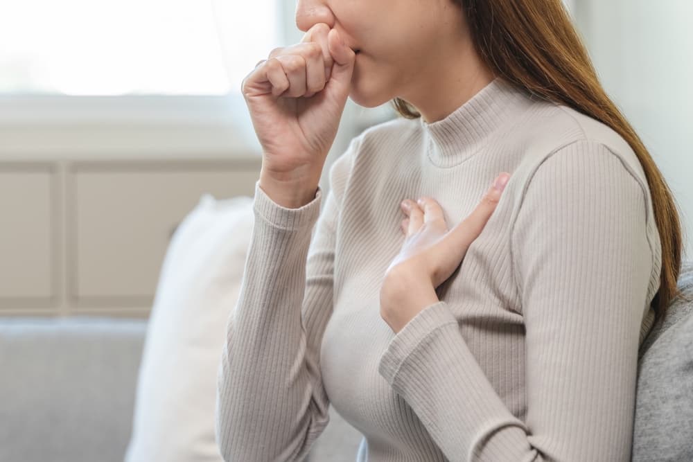 Pessoa aparentando estar com tosse ou algum desconforto respiratório, ela está com uma das mão no peito e a outra na boca, em um gesto tático de tosse.