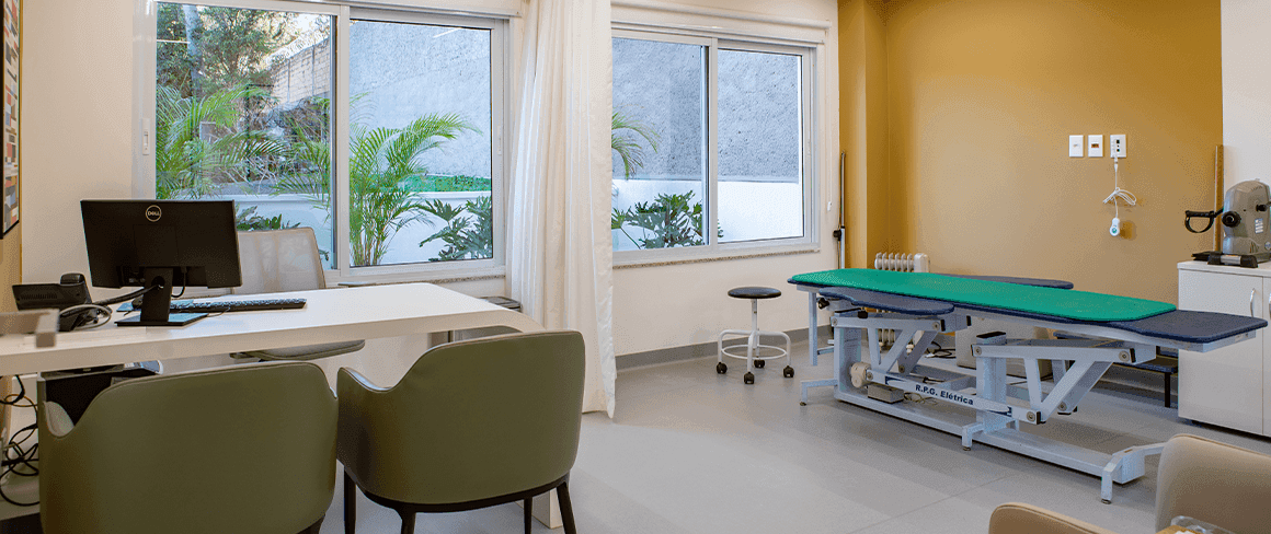 Sala com parede amarela, duas cadeiras e uma mesa, além de uma maca