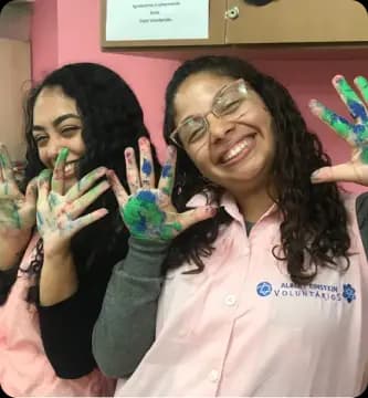 Voluntárias do Einstein sorrindo enquanto mostram mãos pintadas, simboliza a alegria e o impacto positivo do voluntariado, que promove acolhimento e humanização no ambiente hospitalar.