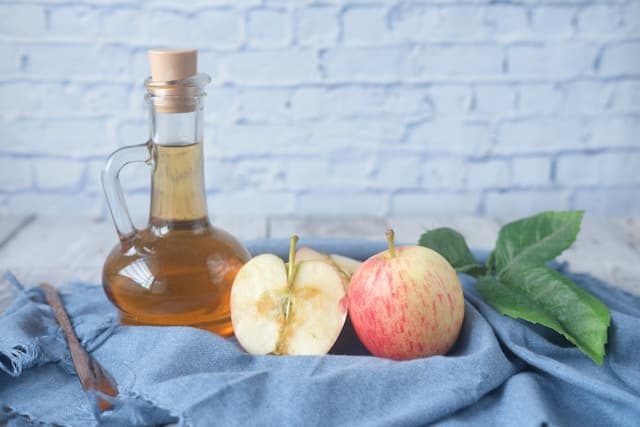 Em uma mesa, uma cesta com um frasco com vinagre de maçã e algumas maças.