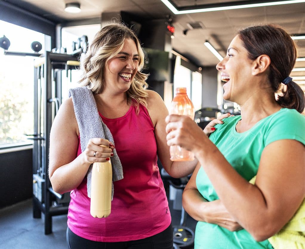 Duas mulheres conversam e sorriem em uma academia, segurando garrafas de água após atividade física, em contexto de saúde e bem-estar da mulher. Duas mulheres conversam e sorriem em uma academia, segurando garrafas de água após atividade física, em contexto de saúde e bem-estar da mulher.