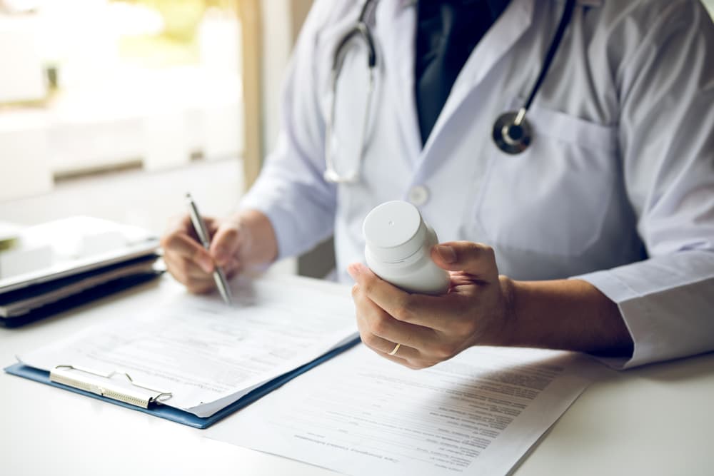 Profissional de saúde com estetoscópio e jaleco com um frasco de medicamento na mão esquerda e com a mão direita fazendo a prescrição para um paciente.