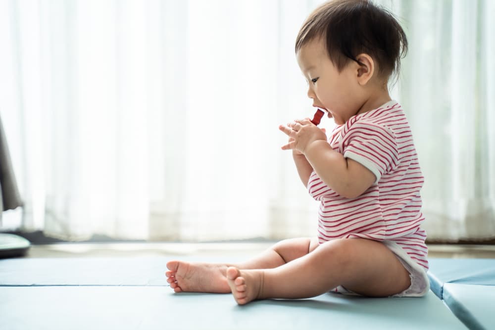Foto com um bebê sentado levando um item pequeno com as mãos até a boca.