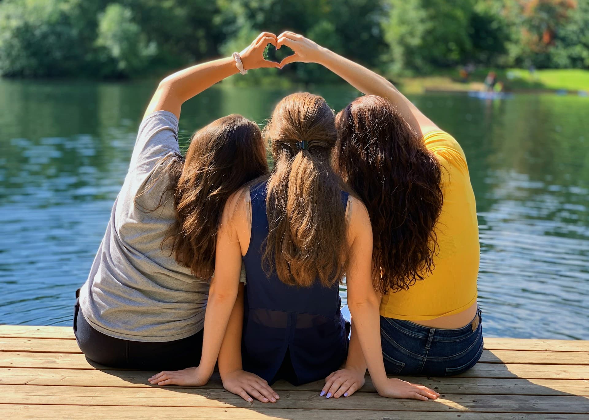 Três jovens sentados em um píer comemorando a amizade com abraços e sinais de coração feitos com as mãos.
