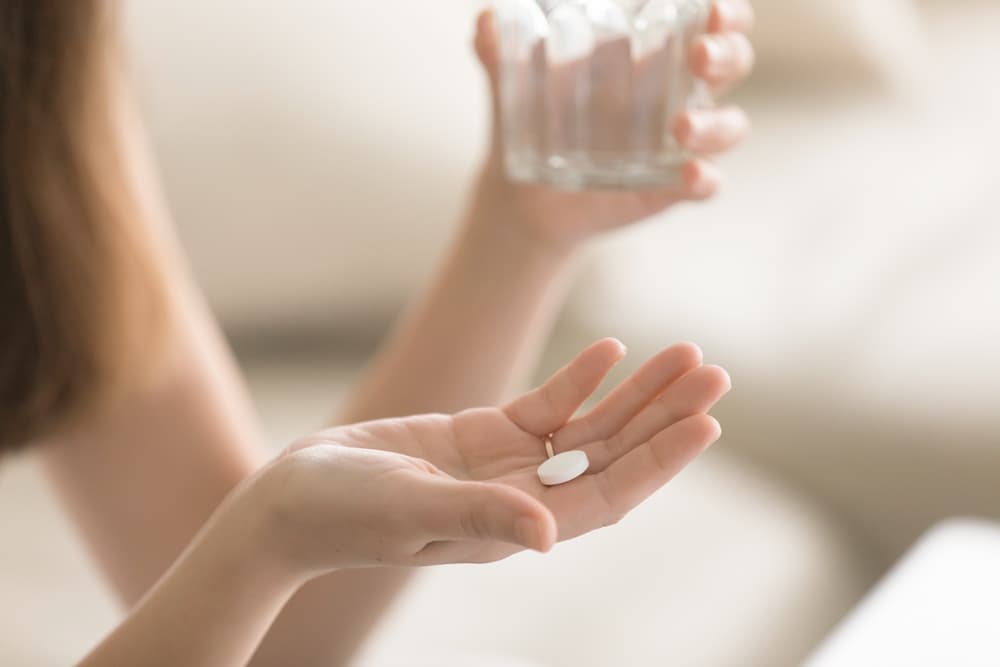 Pessoa com o medicamento na mão direita e um copo com água na mão esquerda.