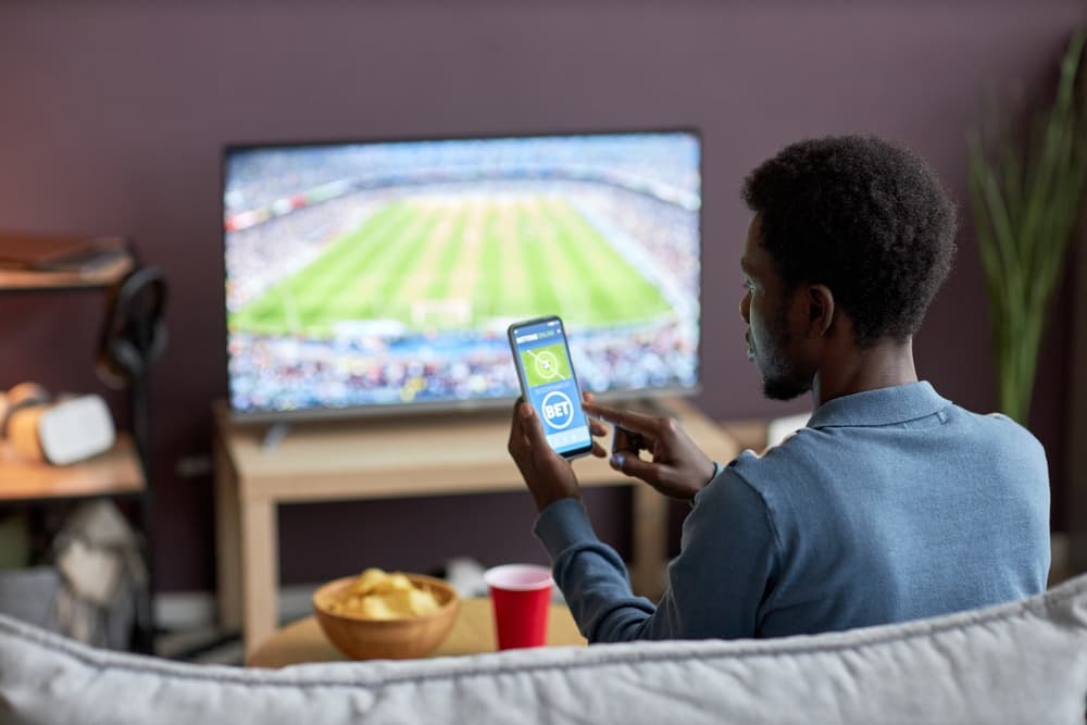Pessoa assistindo a um evento esportivo em uma sala, está sentada em um sofá com um aparelho de celular nas mãos, acessando um site ou aplicativo de apostas - Bet.