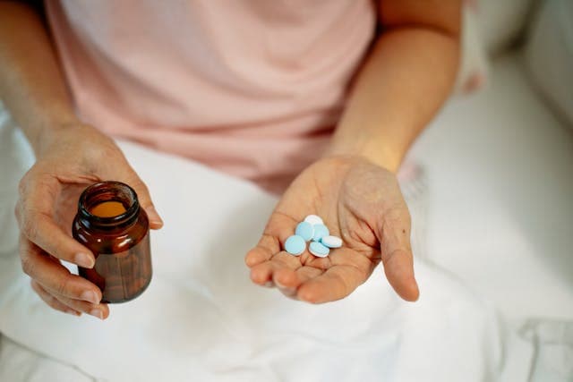 Uma pessoa com um frasco de medicamento na mão direita e comprimidos na mão esquerda está sentada em uma poltrona acolchoada, com um lençol branco sobre as pernas.