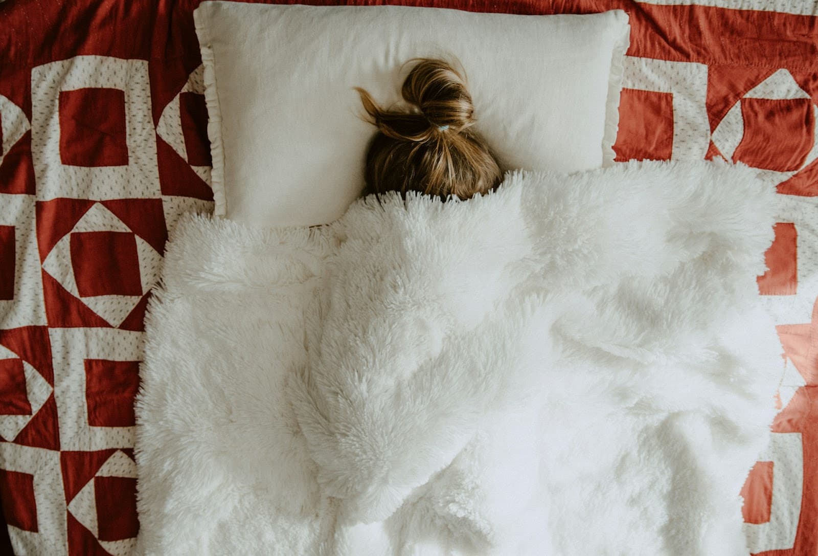 Uma jovem deitada em uma cama com o cobertor sobre o rosto.