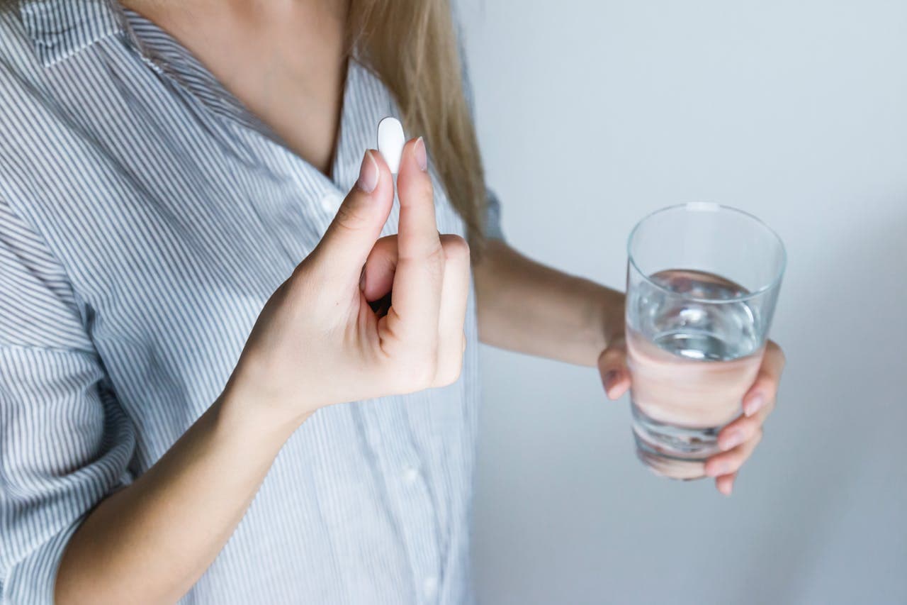 Pessoa com um comprimido na mão direita e, na mão esquerda, um copo com água para se automedicar.