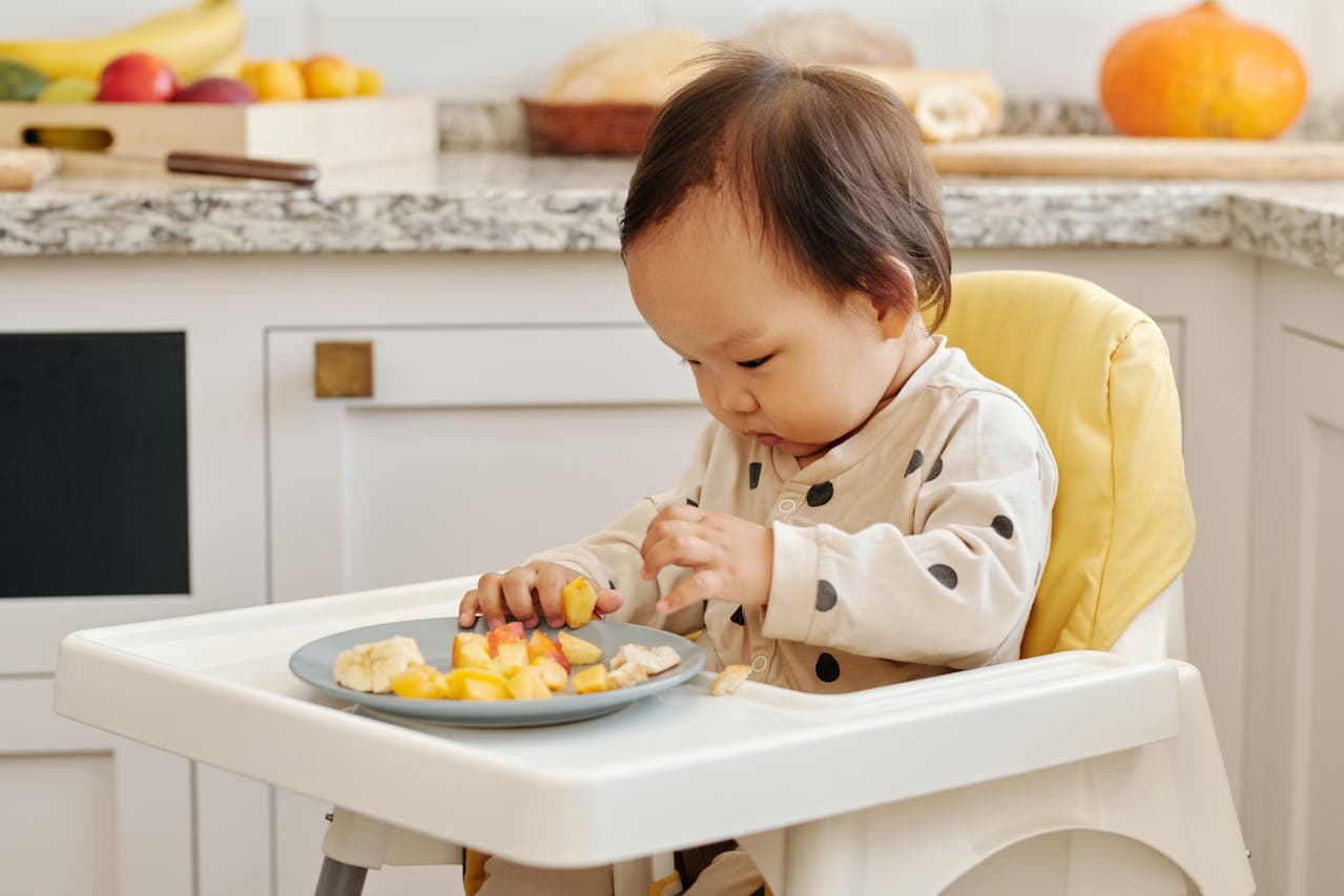 Uma criança sentada em um cadeirão infantil, comendo frutas que estão cortadas e dispostas em um prato.