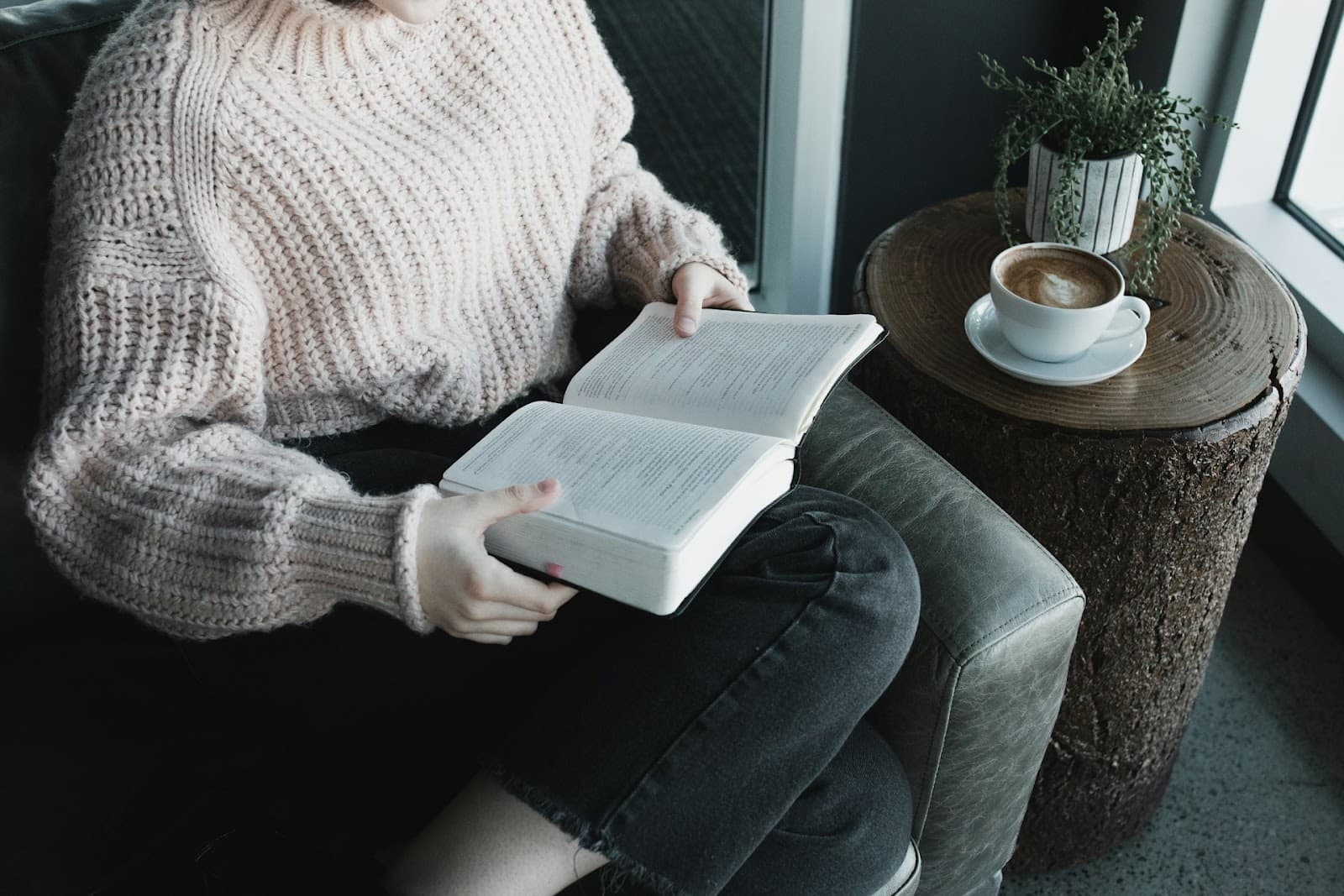 Pessoa sentada em uma poltrona, lendo um livro enquanto saboreia um café.