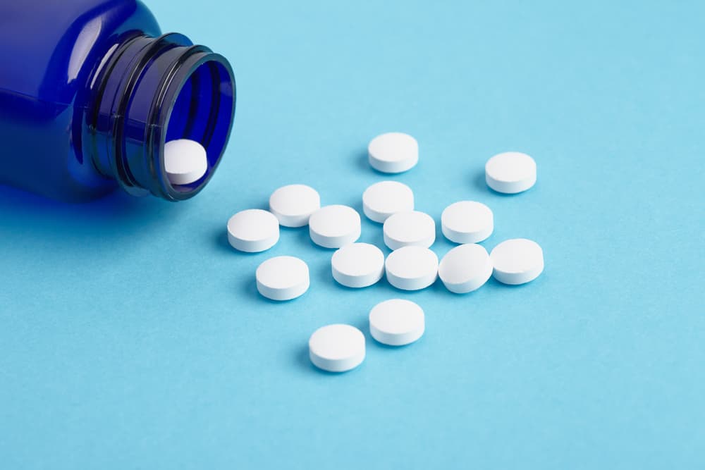 Um frasco azul utilizado para acondicionar medicamentos aberto e comprimidos espalhados sobre uma superfície azul claro.