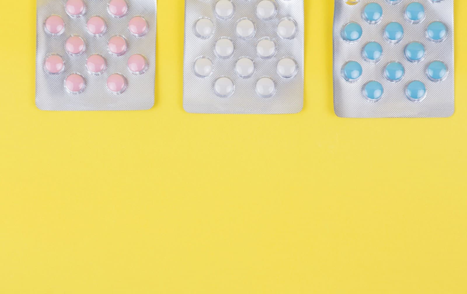 A imagem apresenta três cartelas de comprimidos dispostas em uma superfície amarela. Cada cartela contém comprimidos de cores diferentes. Uma forma ilustrada de diferenciar ibuprofeno e cetoprofeno.