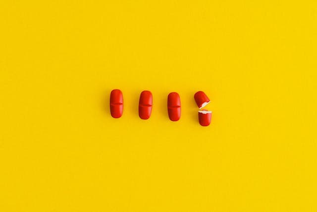 Imagem com fundo amarelo e, sobre ele, quatro comprimidos vermelhos, sendo que um deles está partido ao meio.