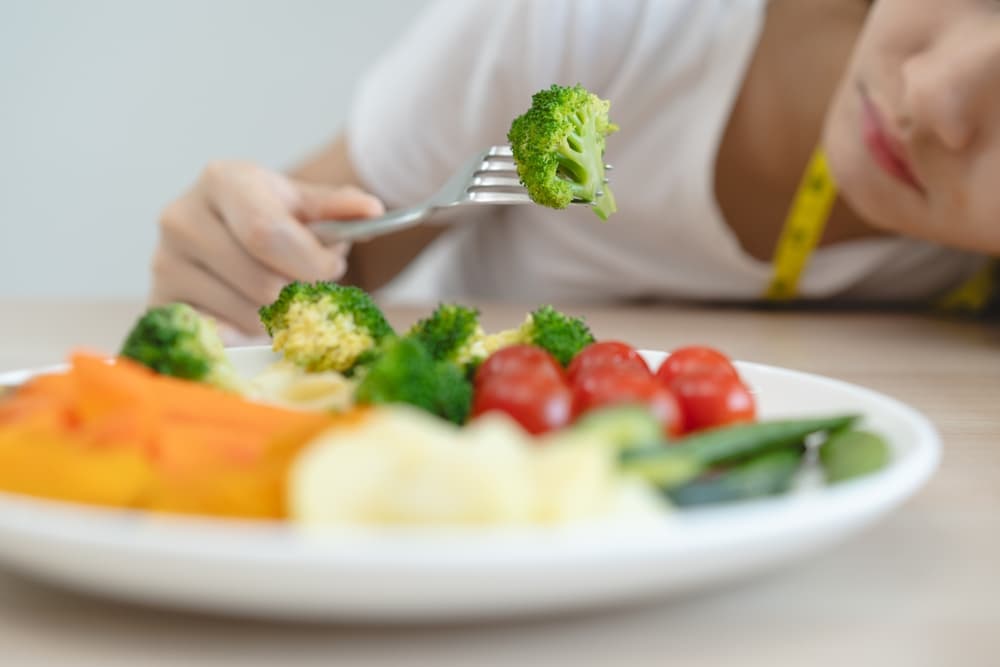 Pessoa sentada à mesa com um prato com uma porção de alimentos e fixamente olhando para um vegetal que acabou de pegar com um talher.