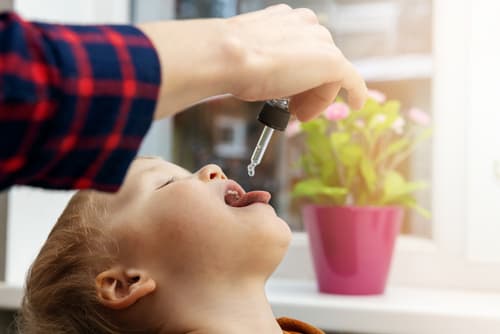 A imagem mostra uma criança recebendo suplementação de vitamina D por meio de gotas administradas com um conta-gotas. Uma mão adulta segura o instrumento enquanto a criança está com a cabeça inclinada para trás e a boca aberta.